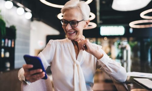 Smiling elderly businesswoman looking at screen of mobile phone while sitting at table with hand touching chin and happy to find positive information on project progress