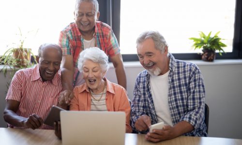 Happy senior people using technology while sitting at table against window in nursing home